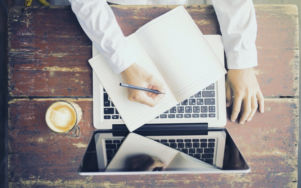 An open notebook and a laptop side by side on a worn wooden desk, a hand writing