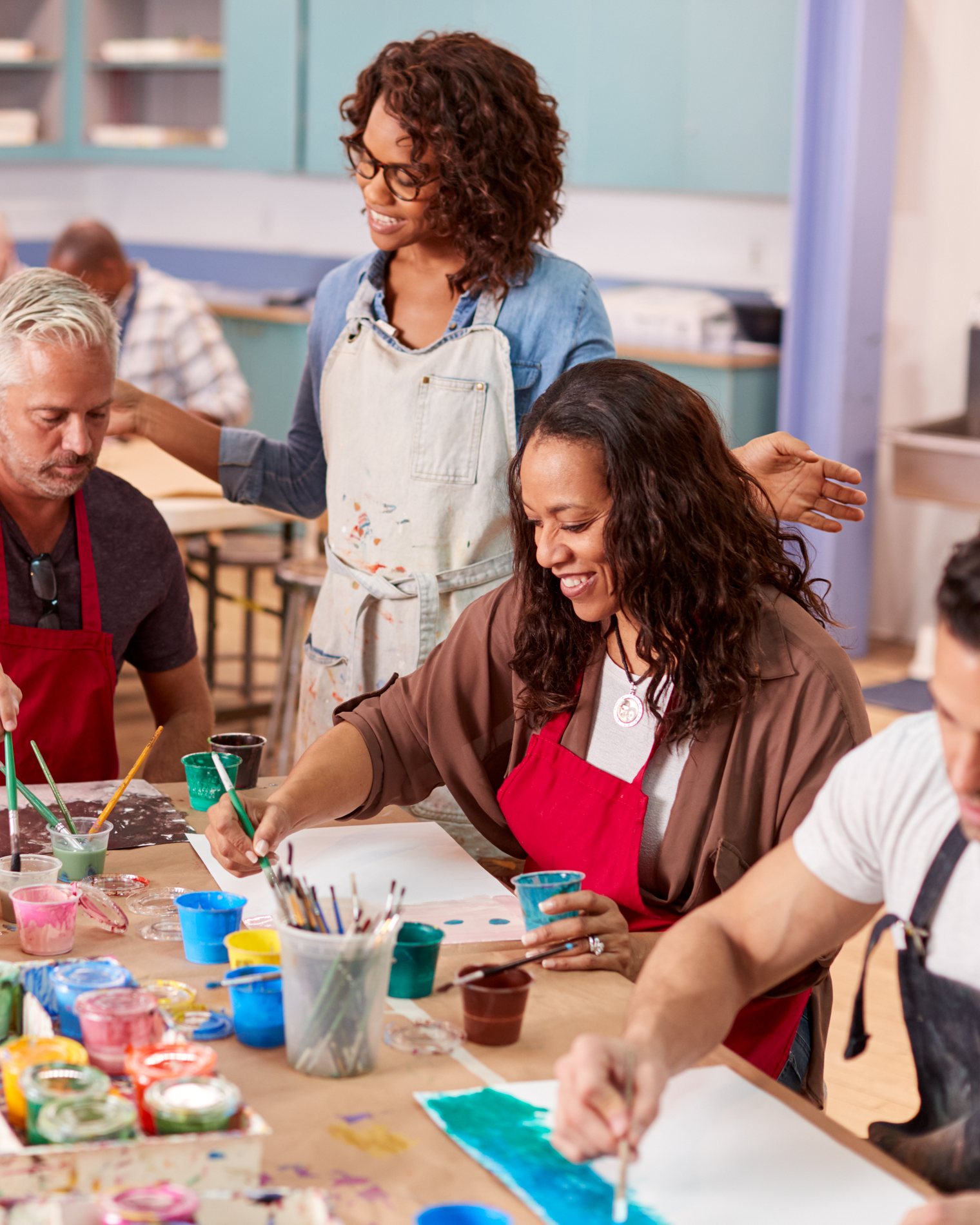 Adults painting at shared tables during a community art class while an instructor looks on.
