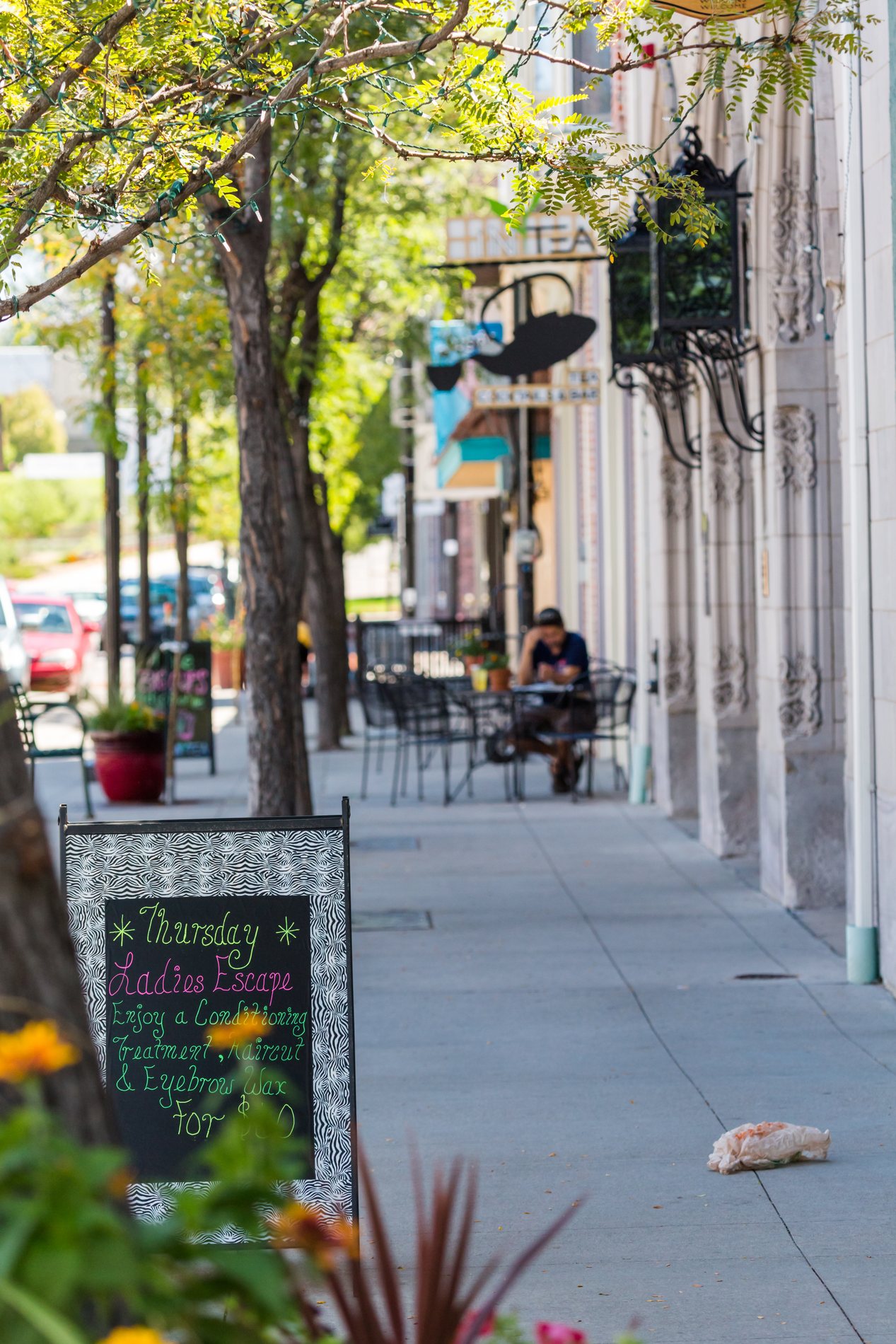 The tree-lined sidewalk of Main Street in Downtown Littleton, with café tables and hanging lanterns.
