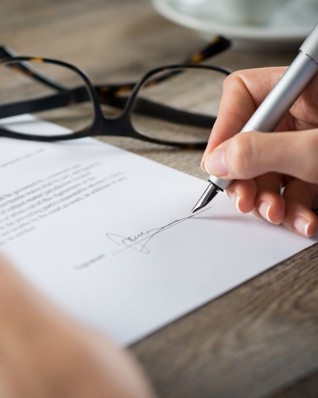 A hand signing an agreement at a desk beside a pair of reading glasses